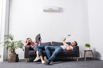 young couple with hand fan and newspaper suffering from heat at home with broken air conditioner