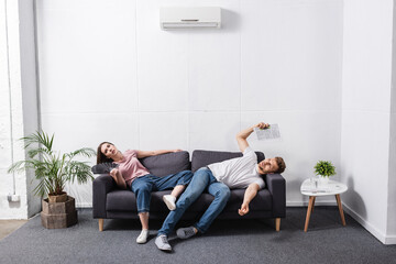 sad couple with hand fan and newspaper suffering from heat at home with broken air conditioner