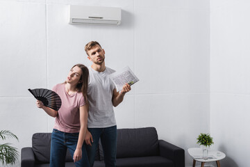 exhausted couple with hand fan and newspaper suffering from heat at home with broken air conditioner