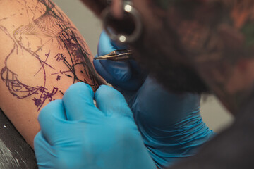 A tattoo artist paints a client's tattoo with ink. Tattoo artist holding a tattoo machine in Blue Sterile Gloves.