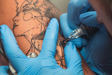A tattoo artist paints a client's tattoo with ink. Tattoo artist holding a tattoo machine in Blue Sterile Gloves.