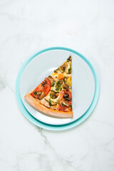 Slice of Vegan vegetable pizza. Dish with tomatoes, eggplant, champignons, Mozzarella cheese and basil pesto sauce. slice on pastel turquoise and gray serving plates. tableware on marble background
