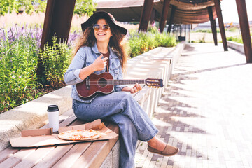 Young pretty woman in hat playing her ukulele outdoors in the modern park and smiling. Carefree mind, concept musical playing, creative activity, music education.