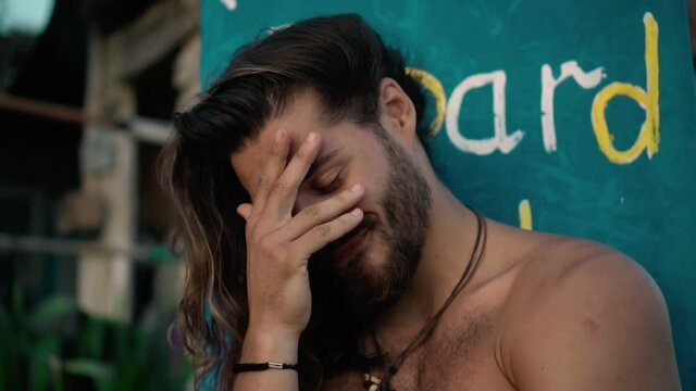 Young handsom European guy brunette with long hair with a wrapped sports body looks forward smiles with an embarrassed look and covers his face with his hand from shame