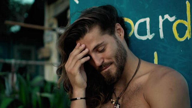 Young handsom European guy brunette with long hair with a wrapped sports body looks forward smiles with an embarrassed look and covers his face with his hand from shame