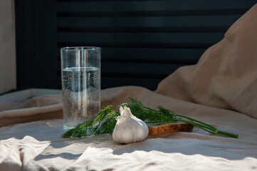 A transparent glass with water, a head of garlic, dill on a slice of bread on a sand-colored linen tablecloth, on a sunny day.