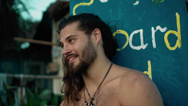 Young handsom European guy brunette with long hair with a wrapped sports body looks forward smiles with an embarrassed look and covers his face with his hand from shame