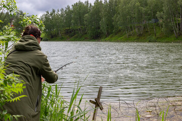 young beautiful fisherman girl fishing with a rod in a large pond at the rain. the woman is wearing the green rain cover. Active sports and relaxation for women