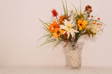 A glass vase with colorful flowers in it. Placed on top of a white wood table next to a white wall. Taken inside with studio bright lights.