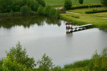 A view of a small lake or pond surrounded from all sides with dense forests, moors, and farmlands with a small wooden jetty or marina with a small platform leading to it located next to the coast