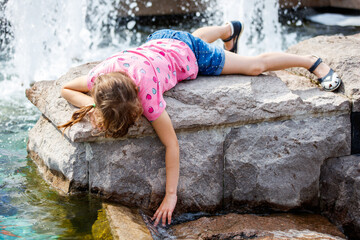 A little girl lies at the fountain and touches the water with her hand