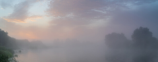 Tranquil morning landscape panorama at sunrise. Heavy fog over the river. Dawn illuminates and makes the clouds colorful.
