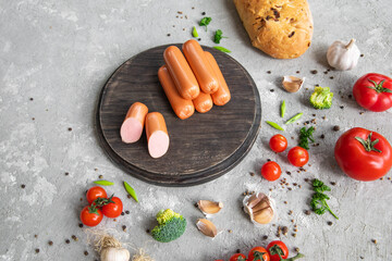 Sausages on dark cutting board with beautiful vegetables, spices and bread. Copy space. Top view. Gray textured background.