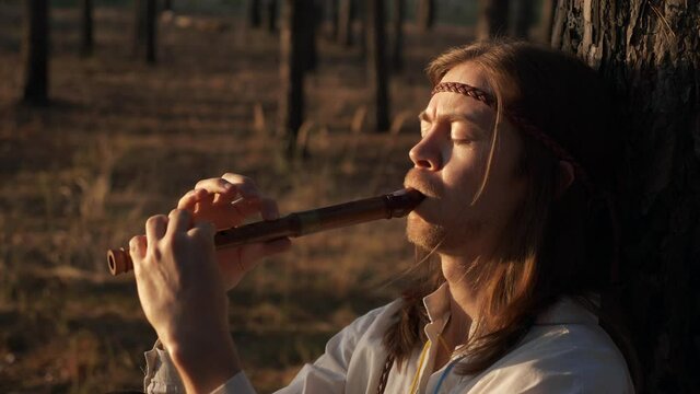 Close-up Man with Long Hair Plays Flute under Pine Trees. On Background Woman with Wreath and Basket Walks in Forest Nature. Slow motion 60 FPS