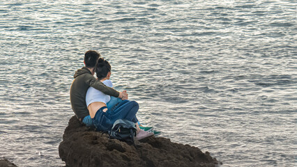 A young man and a girl are sitting on a sea reef embracing and looking at the sea..copy space