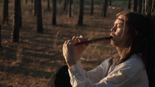 Close-up Man with Long Hair Plays Flute under Pine Trees. On Background Woman with Wreath and Basket Walks in Forest Nature. Slow motion 60 FPS
