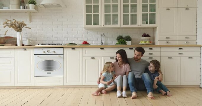 Happy family mom dad and little cute children siblings talking laughing embracing sit on kitchen floor, smiling young parents having fun playing with funny small kids bonding together at home
