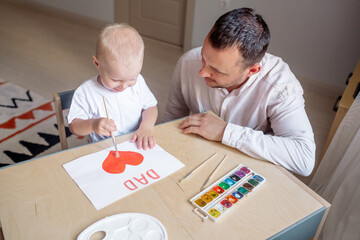 Little boy 2 years old makes a card for the holiday Father's Day with his bearded young father