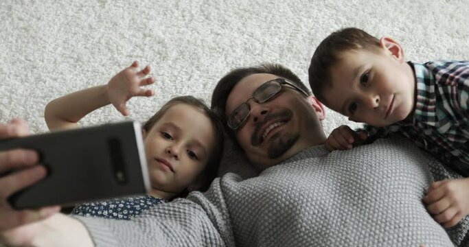 The family lies on the floor and takes a selfie. Dad with son and daughter are talking on a video call on a mobile phone.