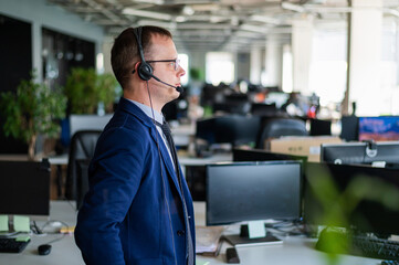 Portrait of a male call center operator in headset at workplace. A man works in an open space office answering customer calls. Support service or hotline. Agent of a telemarketing company.
