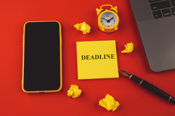 Alarm clock as time passing concept for business deadline, urgency and running out of time. Deadline message on yellow sticky note. Smartphone, alarm clock and laptop on red background.