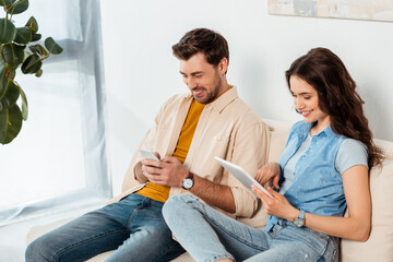 Smiling couple using smartphone and digital tablet in living room