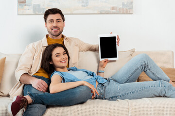 Young couple smiling at camera while showing digital tablet with blank screen in living room