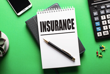 The word INSURANCE is written in a white notebook on a green table near a calculator and a phone. Insurance concept
