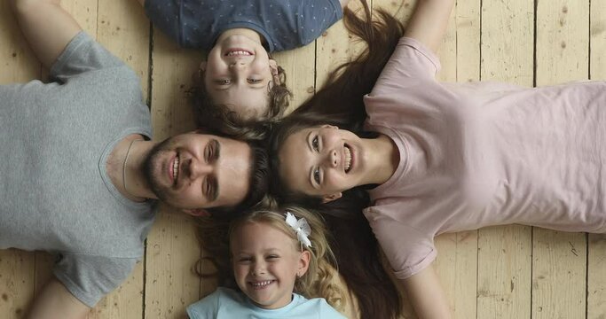 Happy parents and cute little kids son daughter lying in circle on warm wooden floor, smiling dad mom and children bonding looking at camera, family portrait, underfloor heating concept, top view