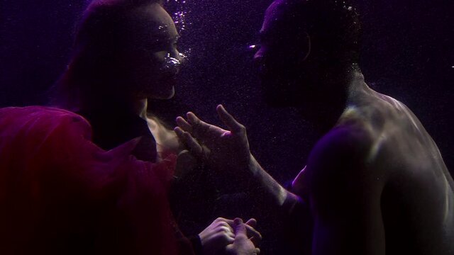 Close-up portrait of a tender couple in love who are under water and look at each other, a buxom girl in a dress and a dark-skinned man. They are on a dark background with bubbles.
