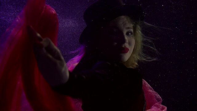 Close-up portrait of a girl in a black hat and red lipstick on her lips, she is under water. It floats on a dark background with the fabric from the skirt fluttering around it.