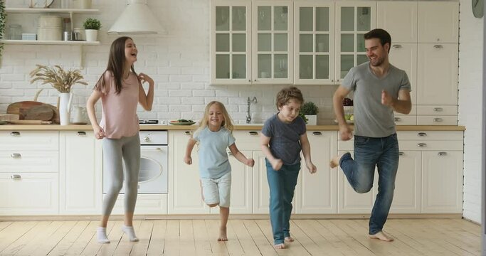 Happy active family young fit sporty parents and two cute small children jumping together in kitchen, carefree funny little kids with father mother having fun do morning exercises laughing at home