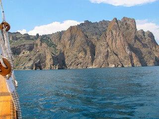 Naklejka premium A picturesque panorama of the impregnable cliffs of the extinct volcano Karadag from the side of a pleasure yacht against a bright blue sky with white clouds and blue sea