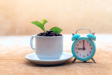 Roasted coffee beans In a white coffee cup has green leaves and an alarm clock and on a light brown fabric background and a few coffee beans scattered.
