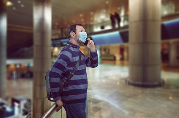 Mature man in medical face mask leaning on a railing in an airport’s half-empty transit hall. He is talking on a cell phone.