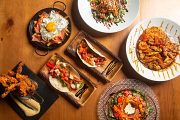 Varied food dishes of the world on wooden table, eggs, salad, chicken, tacos.