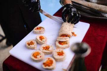 A cook with black gloves on a board cuts a roll of white meat stuffed with vegetables