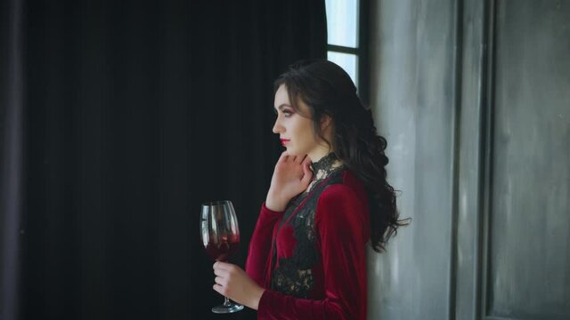 Young beautiful sexy fairytale mystical vampire queen. Woman lady holds glass bloody wine in hand. Brunette black long wavy hair. Red velvet masquerade vintage dress. Dark interior gothic room, window