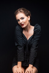 Caucasian attractive sexy professional female model with brown hair bites her lip and poses in the studio in a dark dress, sitting on a chair, on black background