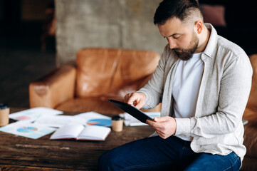 Young bearded guy in stylish casual clothes uses his tablet while sitting on a table in his creative office