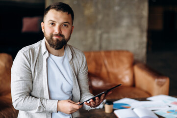 Attractive young  guy with of Caucasian ethnicity sits in his modern office alone, holding a tablet in his hands and smiling