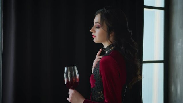 Artwork. Young beautiful woman posing image fabulous angry witch vampire queen. Girl gently touches neck black necklace. Holds glass bloody wine in hand. Moves head, lips, closes eyes, agrees says yes