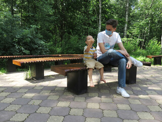 Fototapeta premium Father and son in the park, sitting on a bench and drinking some water. Man in a medical mask on a walk. 