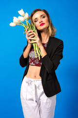Young sensual beautiful woman holding bouquet of white tulips and closing her eyes, smart casual outfit, blue studio background.