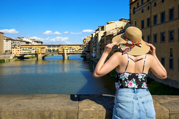 Fototapeta na wymiar Beautiful Young Tourist Girl wearing Floral Dress and Big Hat Walking on Ponte Alle Grazie Bridge in Florence with Ponte Vecchio Bridge in the Background