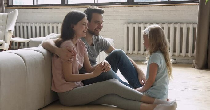 Happy family mom dad and little cute child daughter talking laughing embracing sit on floor, loving young parents having fun with funny small kid girl playing chatting hugging together at home