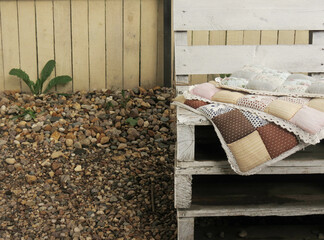 Soft pillows with floral print on a white wooden bench on the street, in the background a green dandelion and stones.