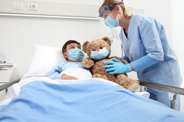 nurse takes care of the patient child in hospital bed playing with teddy bear, wearing protective masks, corona virus covid 19 protection concept,