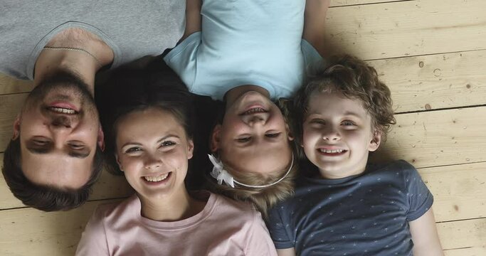 Happy family of four with children laughing looking at camera lying on warm wooden floor, young parents and cute little kids son daughter smiling faces bonding together, portrait, top view from above