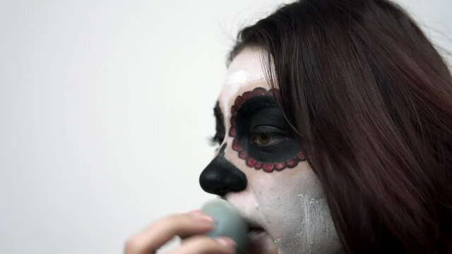 Woman applies white paint to her face with sponge for santa muerte makeup. The girl is sitting in the studio and draws sugar skull at her face. Theme of death day and it's traditional makeup.